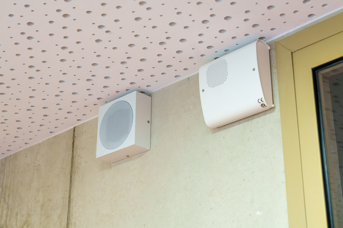 Deux boîtiers sonores blancs fixés en hauteur sur un mur, près d’une porte.