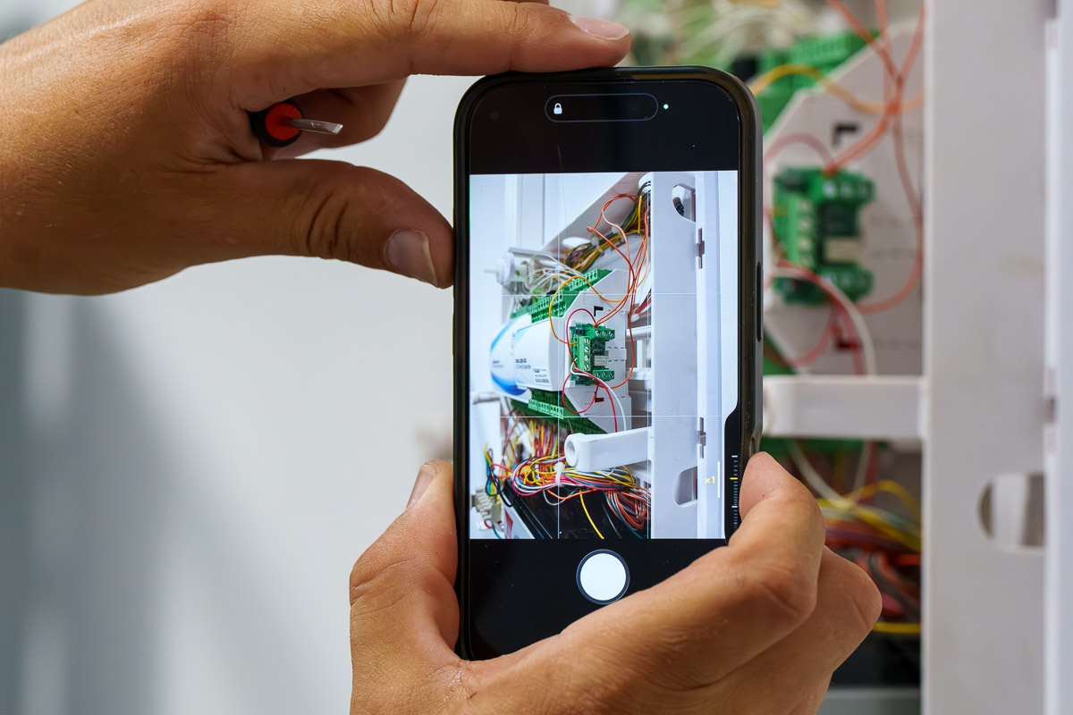 Technicien photographiant avec un smartphone le câblage d’une centrale d’alarme ouverte.